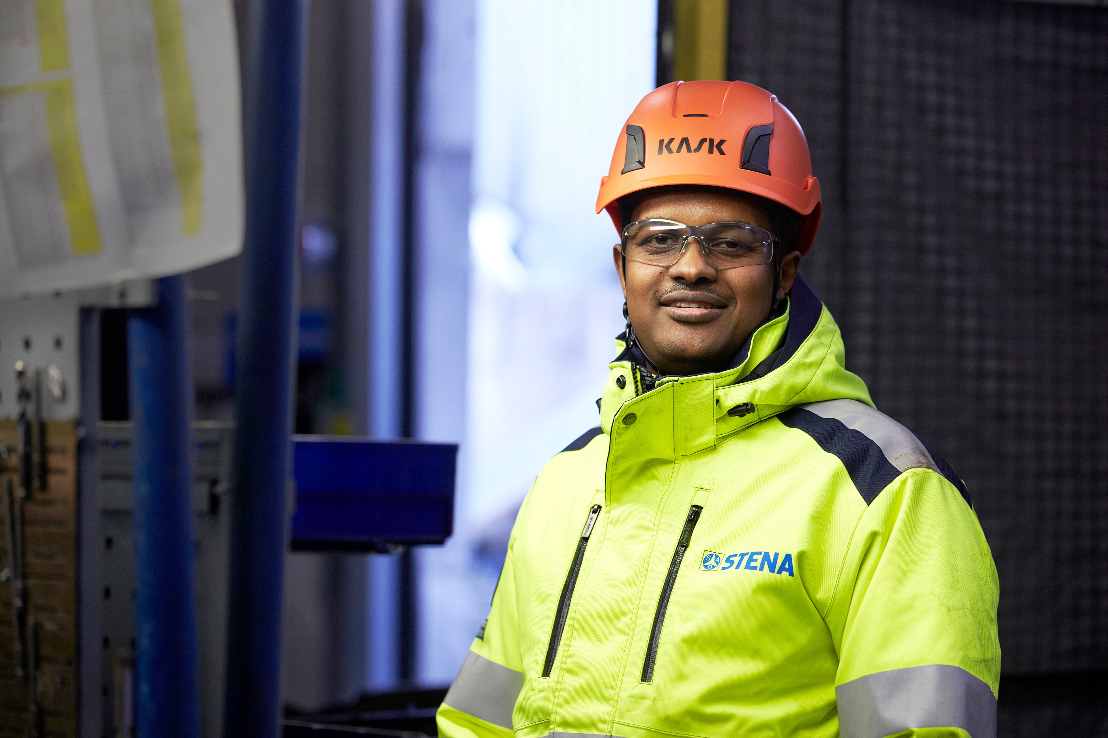 Male Stena Metall employee in hi-vis protective gear and helmet is standing in a facility, smiling and looking into the camera.
