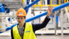 A female Stena Recycling employee in hi-vis protective gear, raising her left hand. In the background, yellow, blue and white machinery is visible.