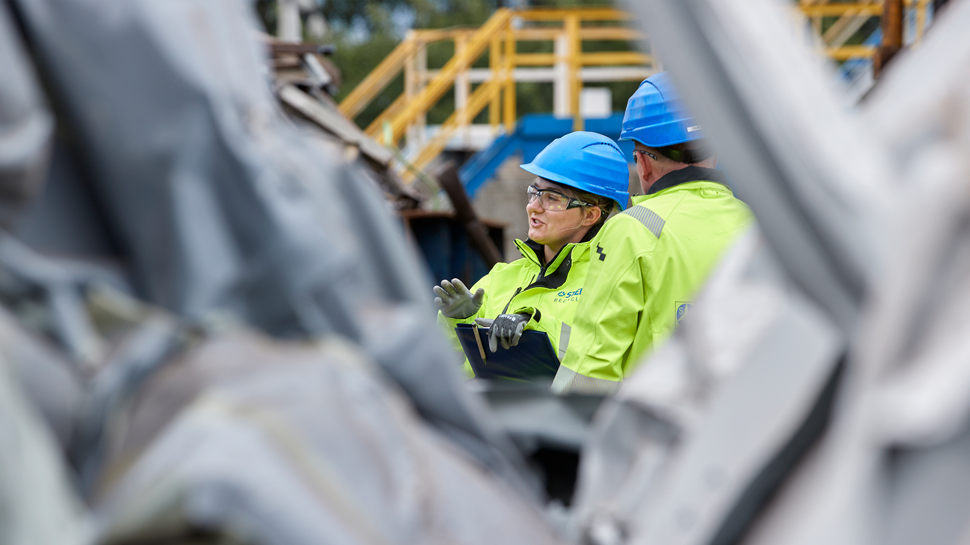A female and a male Stena Recycling employee in high-vis protective gear and helmets stand outside in a recycling facility in Poland where metal among other material is being recycled