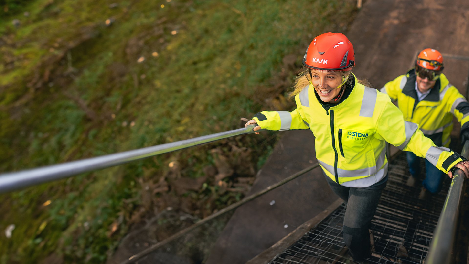 Två Stena Recycling-anställda i varselkläder klättrar uppför trappan utomhus utanför en återvinningsanläggning