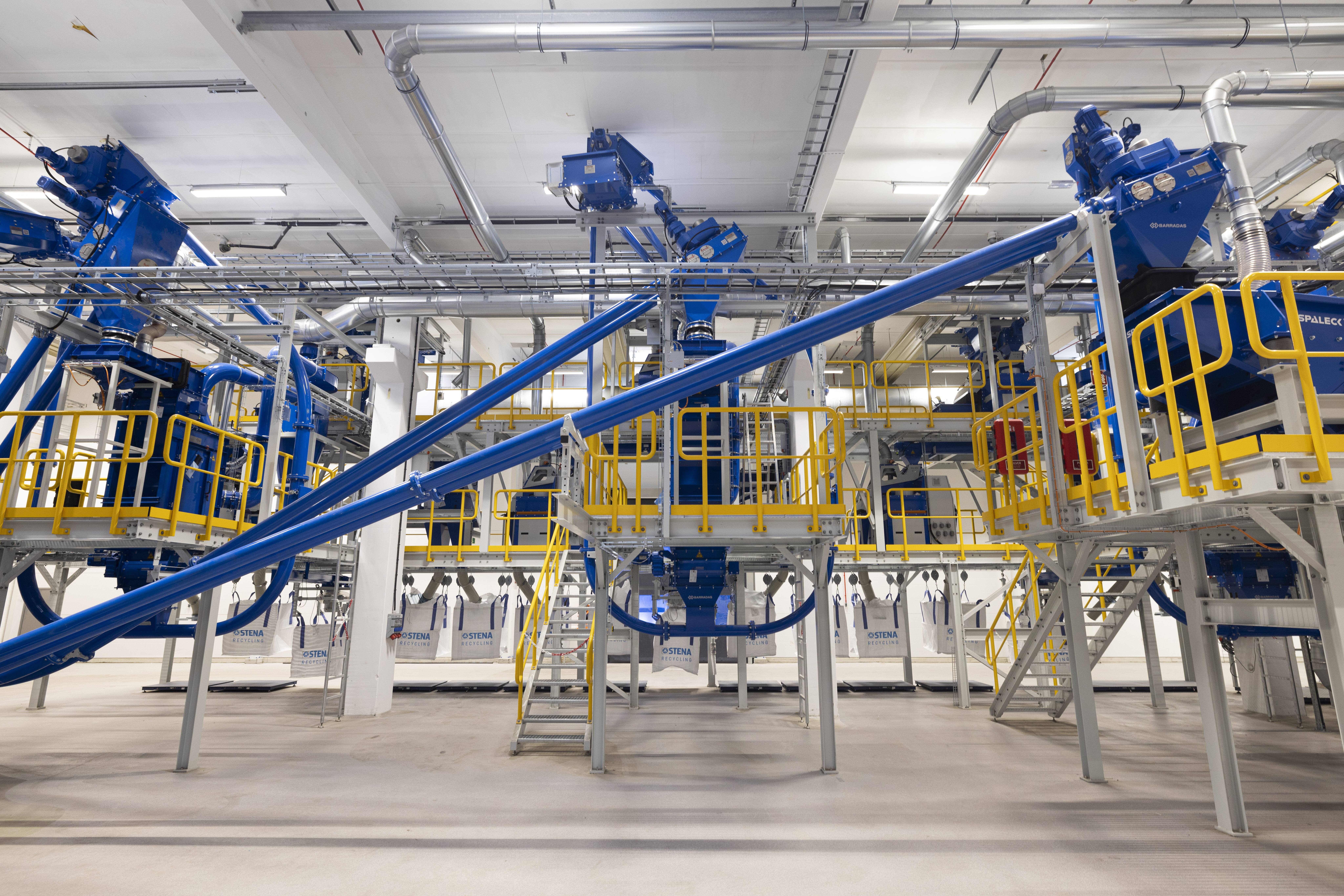 Inne i en återvinningsanläggning. Gula, blå och vita maskiner, rör och stegar.