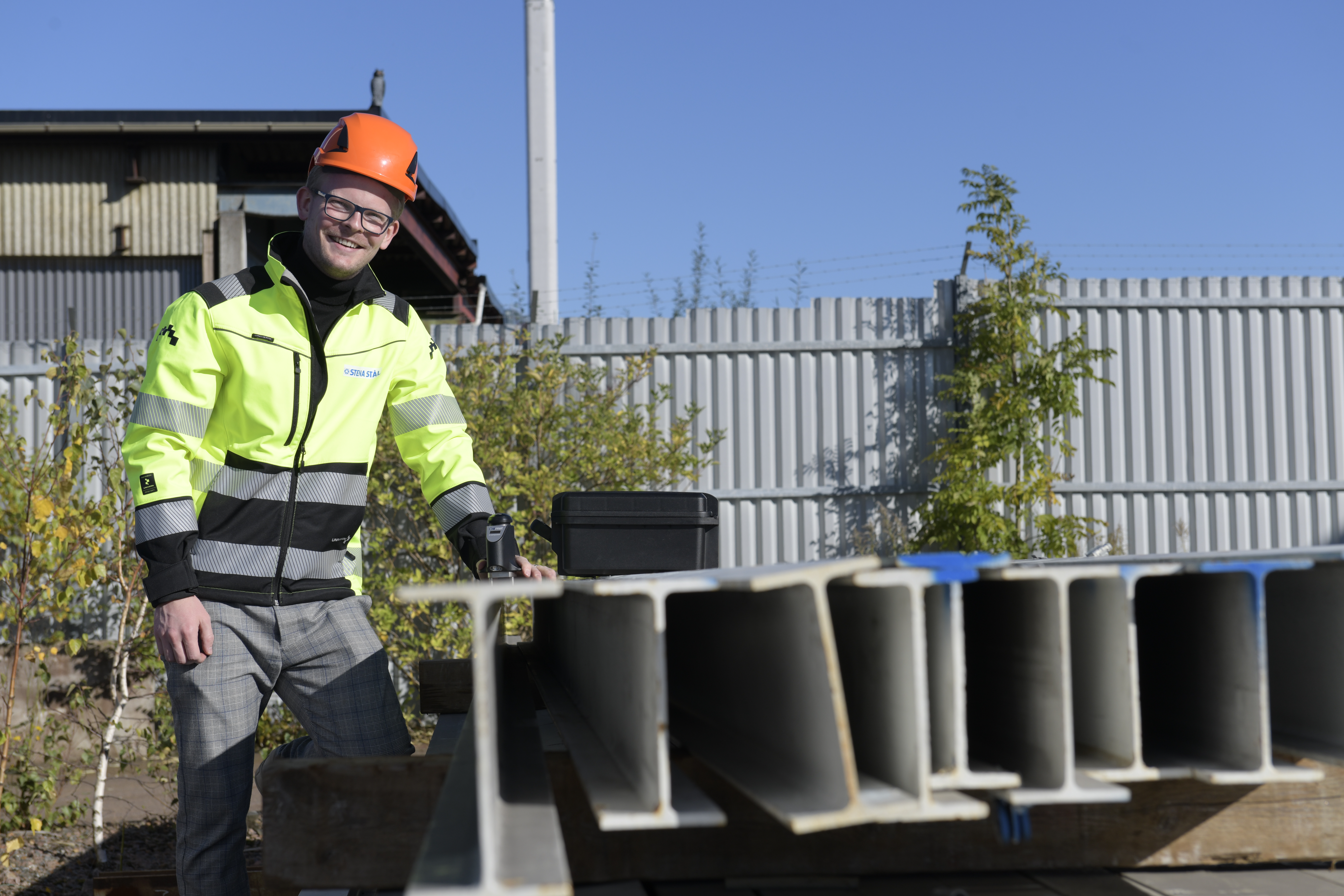A male Stena Stål employee in hi-vis protective gear, standing beside steel beams, with a measurement device in one hand.