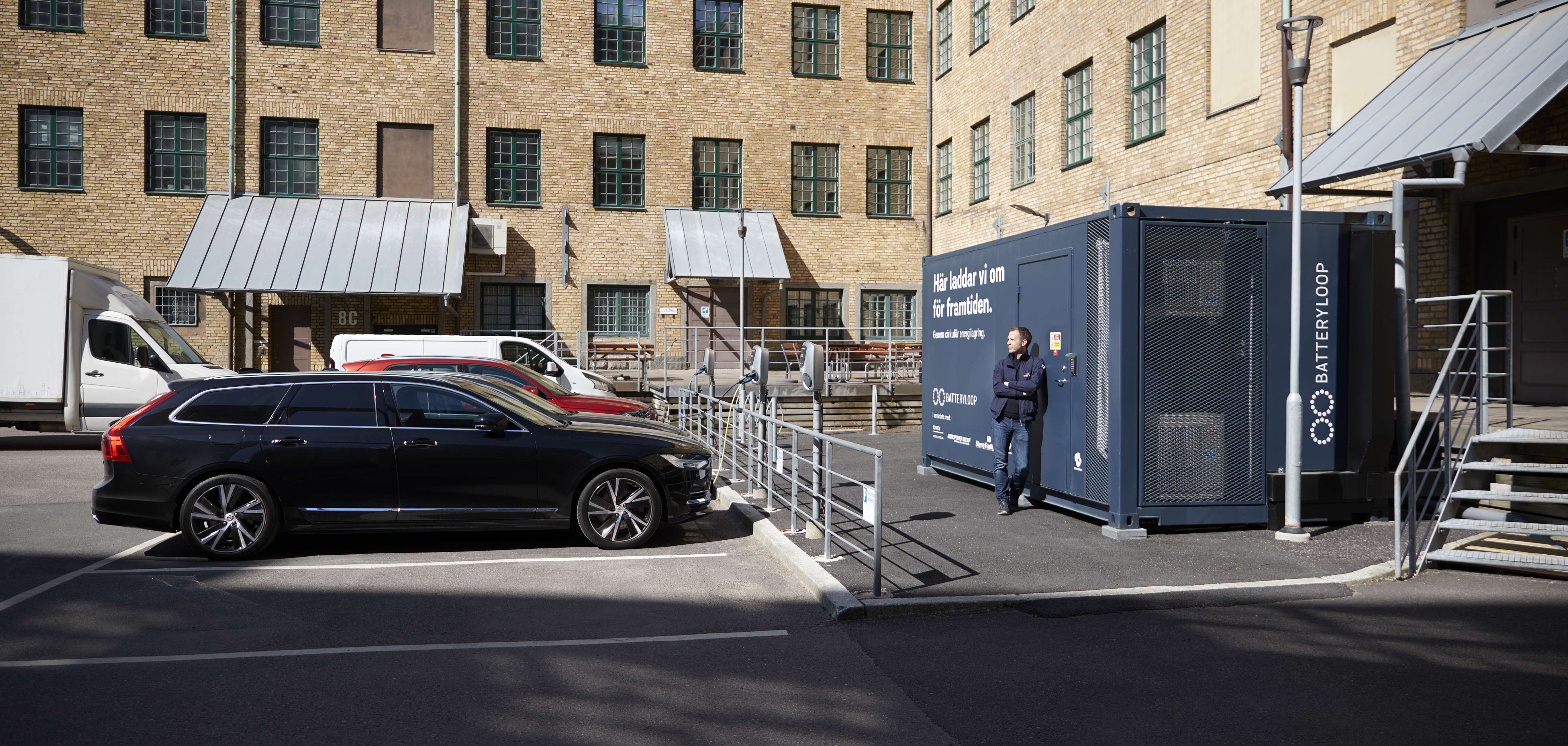 A male batteryLoop employee stands in a courtyard, leaning towards the front of a container. To the left cars, connected to electric chargers.