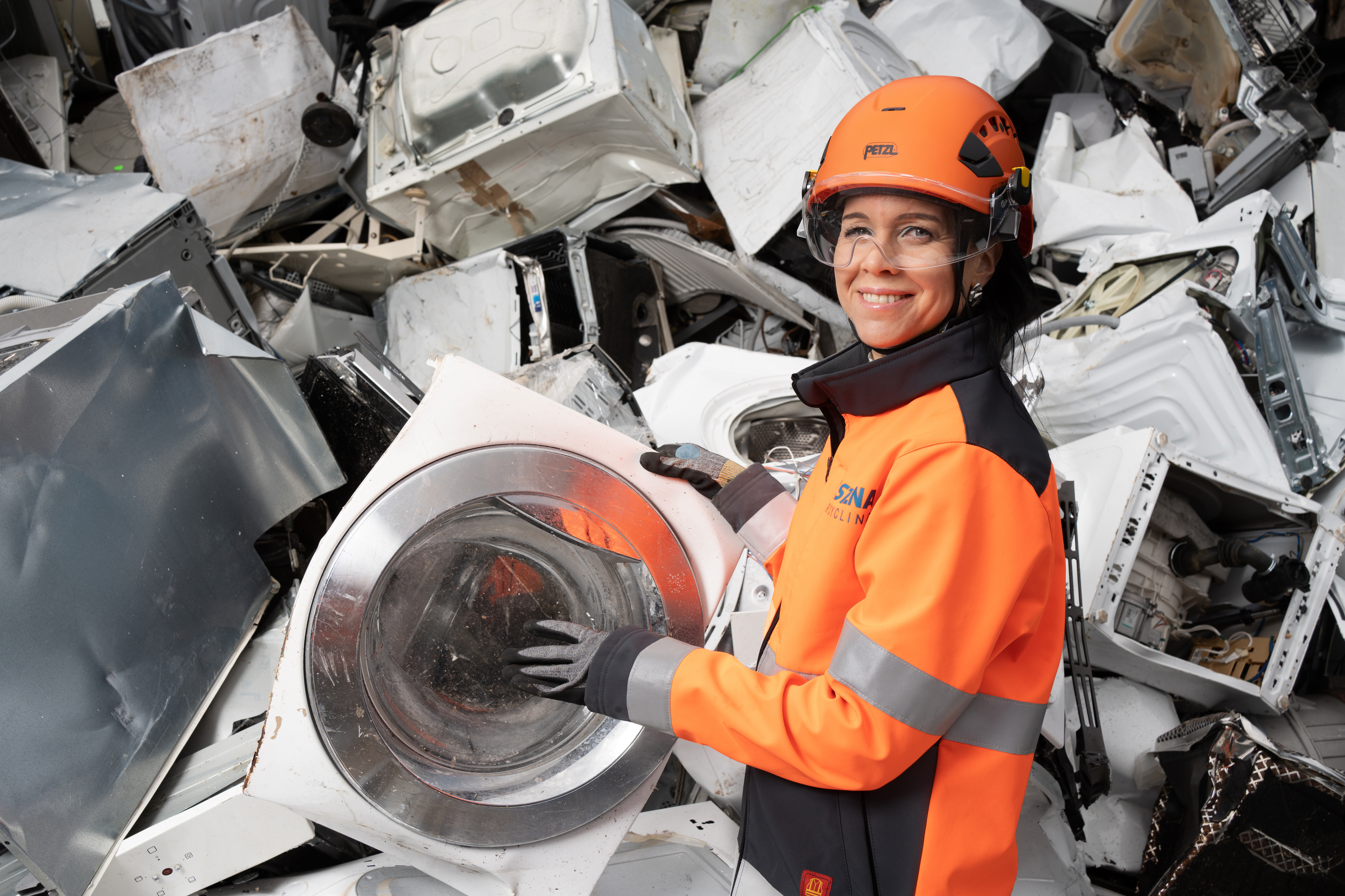 A female Stena Recycling employee in hi-vis protective gear, standing in front of a pile with metal scrap, holding a piece from a washing machine.