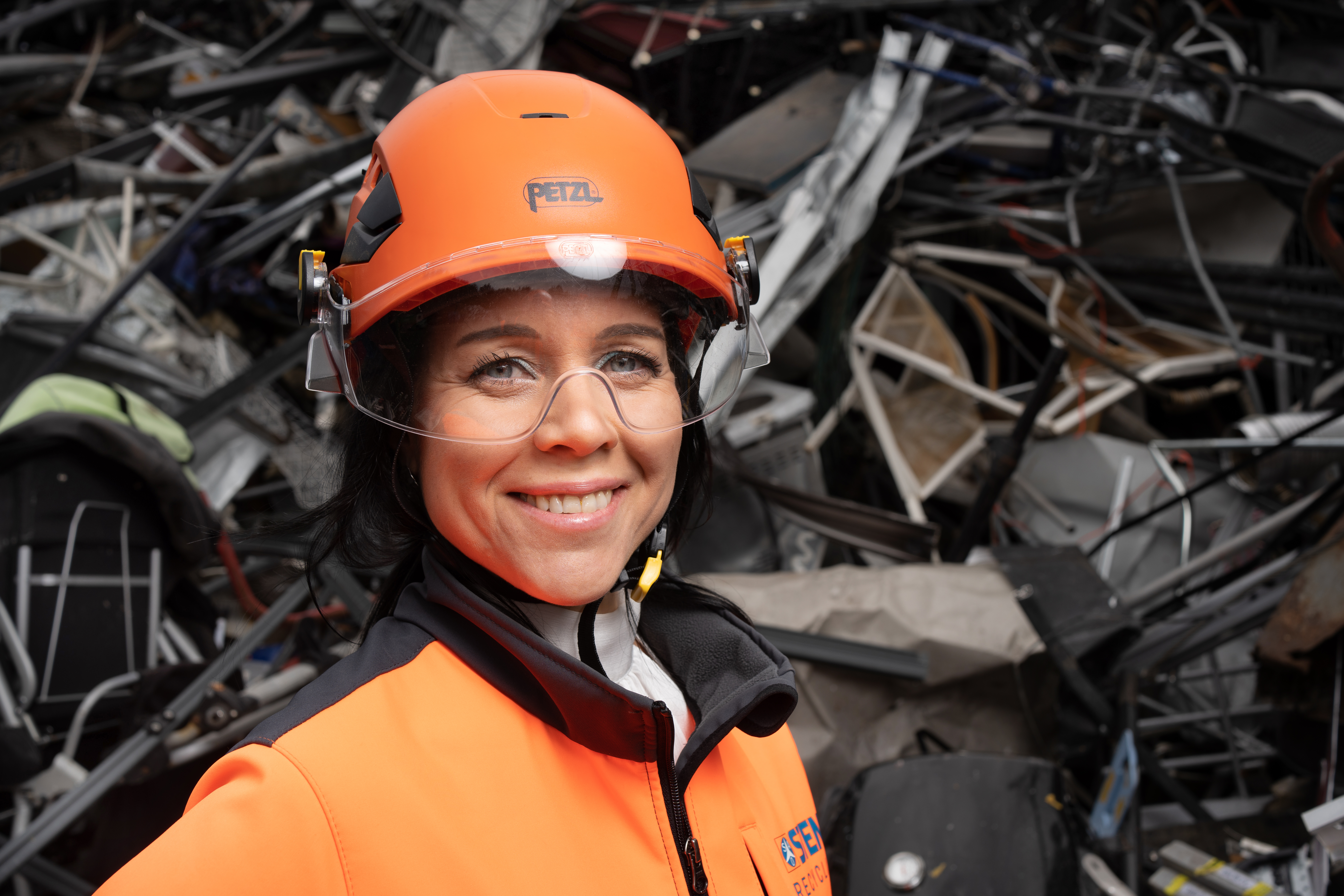 A female Stena Recycling employee in hi-vis protective gear, standing in front of a pile with metal scrap, holding a piece from a washing machine.