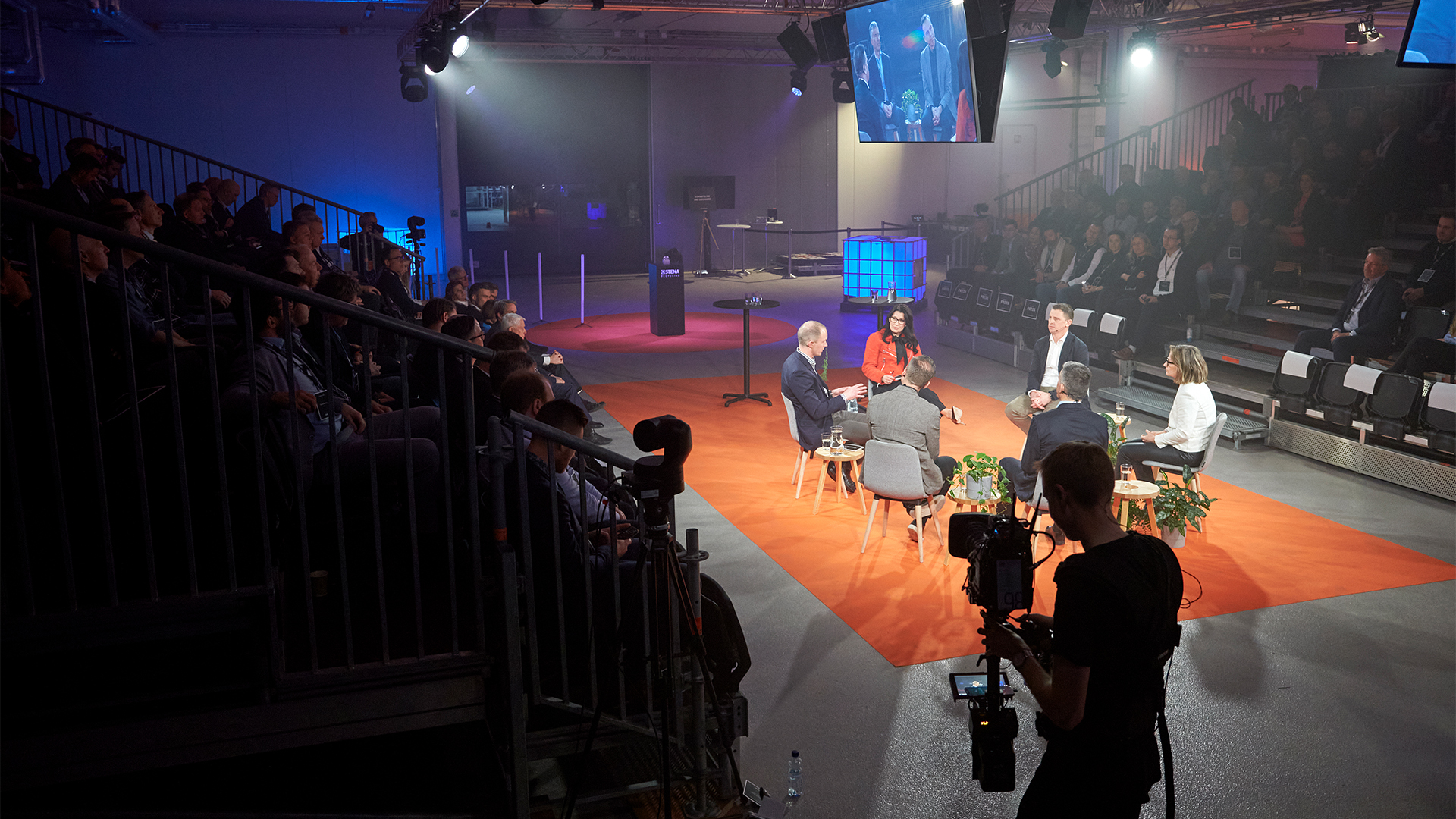 A seminar setting where the audience sits in an amphitheatre-setting. In the middle, a group of people is sitting on chairs, turned to each other.