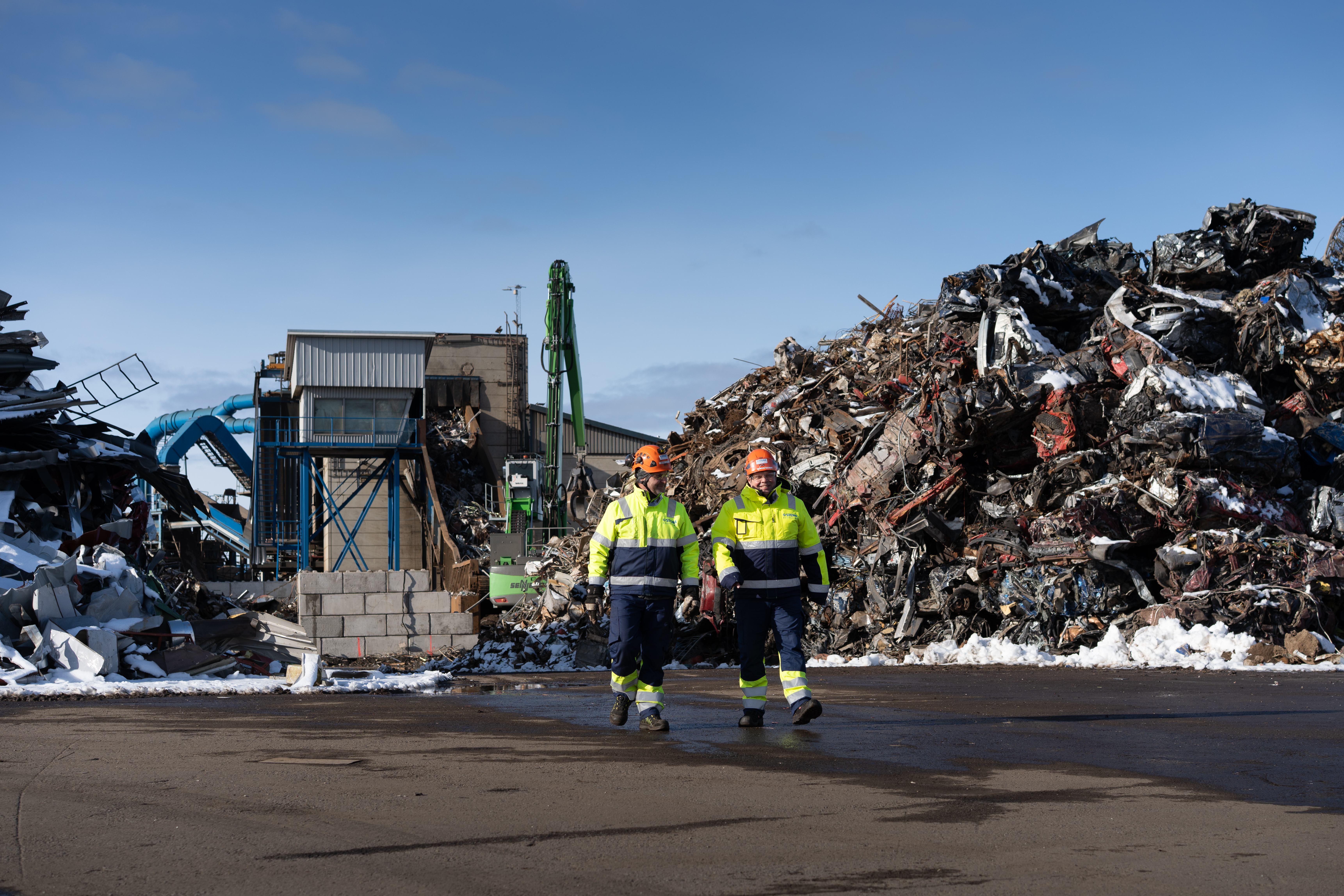 Två manliga anställda på Stena Recycling arbetar utomhus på återvinningsanläggningen i Pori i Finland. Fragmeteringsmaskinen i bakgrunden används för att bearbeta metallskrot till mindre bitar. Arbetet innefattar hantering av tunga maskiner och fordon. Säkerhetsfrågorna står därför högt på dagordningen och berör alla