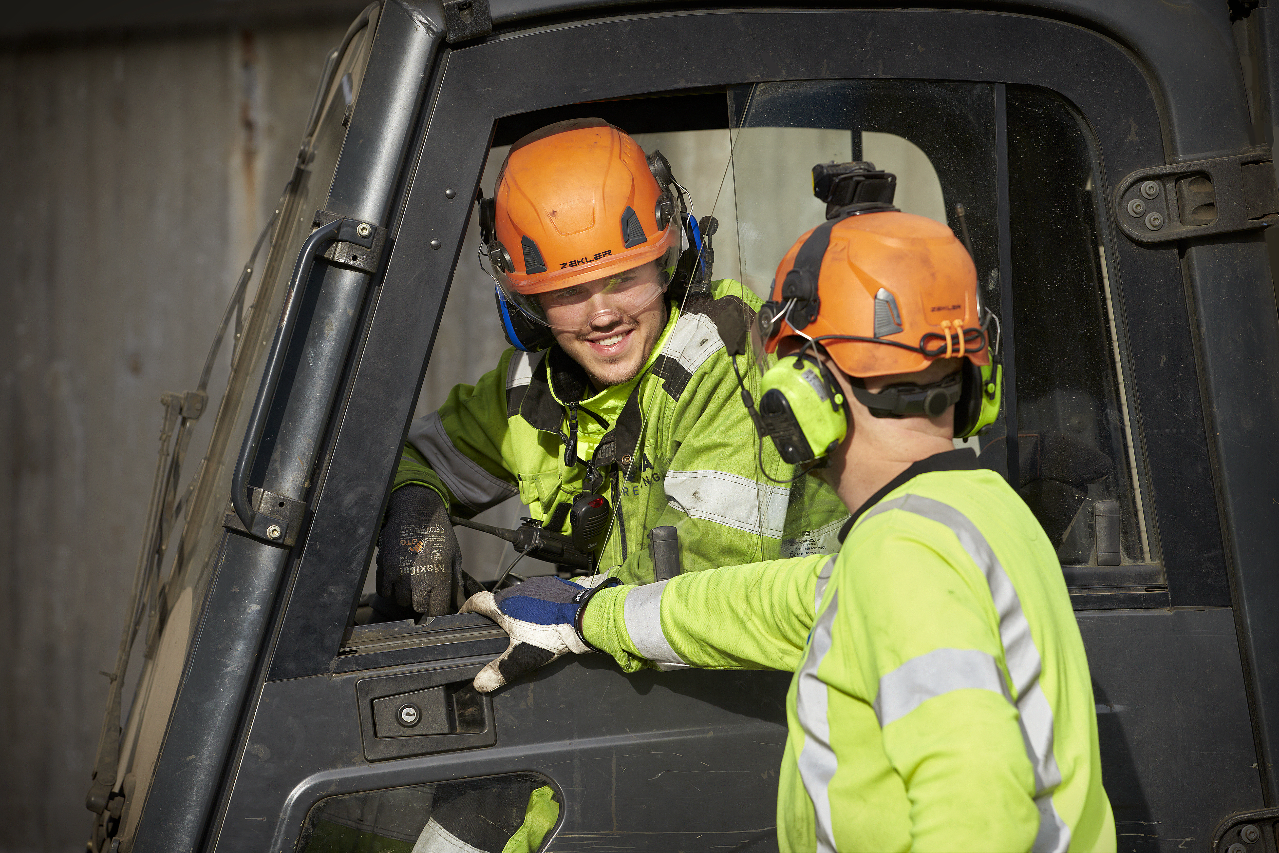 En manlig Stena Recycling-anställd i skyddskläder sitter i förarhytten i en verkstadsmaskin och pratar med en annan manlig Stena Recycling-anställd i skyddskläder.