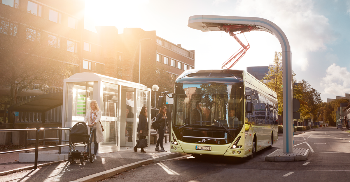 An electric bus has stopped at a bus stop in an urban environment. Two women are about to go onboard the bus. A woman with a stroller passes by on the sidewalk.