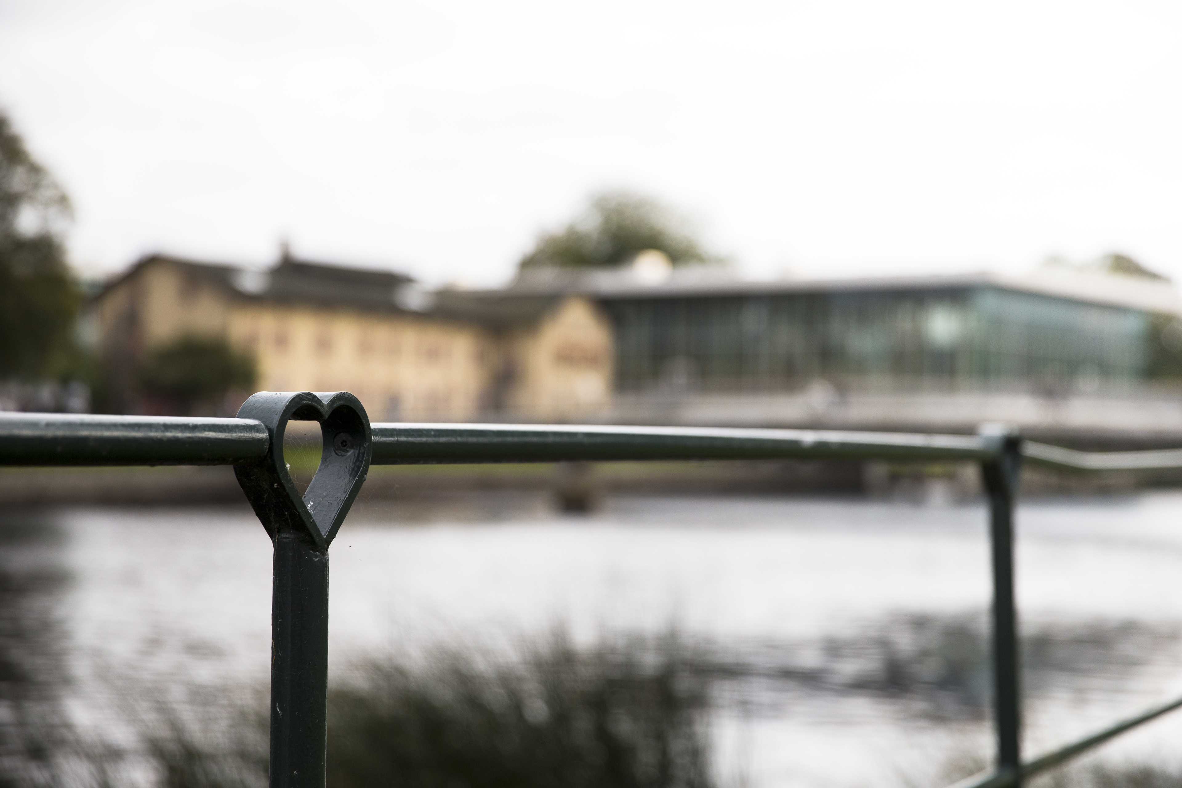 En hjärtformad ledstång på en bro med en suddig byggnad i bakgrunden över floden.