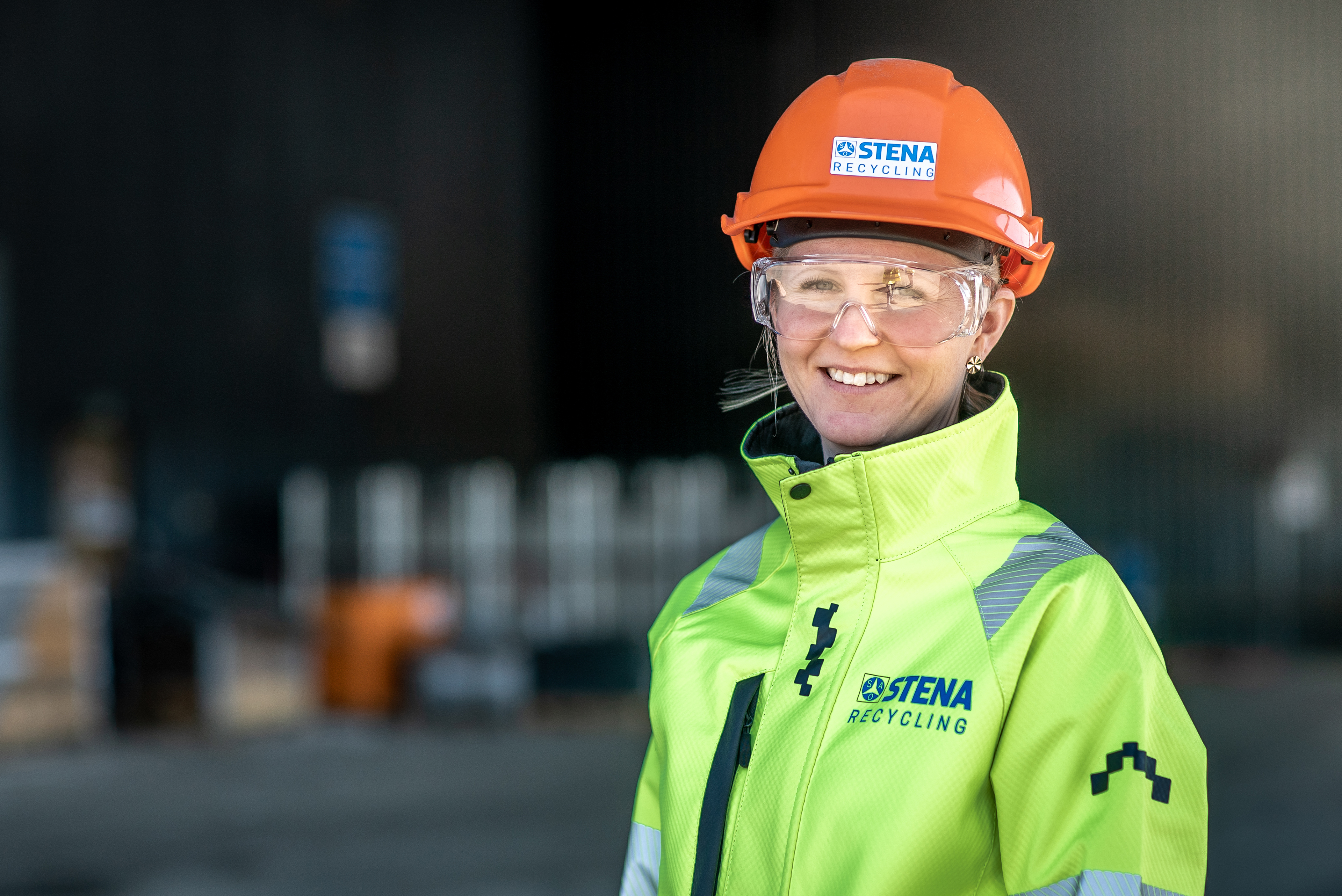 Kvinnlig expert från Stena Metall Group som tittar in i kameran, klädd i reflekterande arbetsjacka och hjälm med Stena Recyclings logotyp