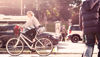 En kvinnlig cyklist cyklar genom staden med bilar, buss och fotgängare i bakgrunden. 