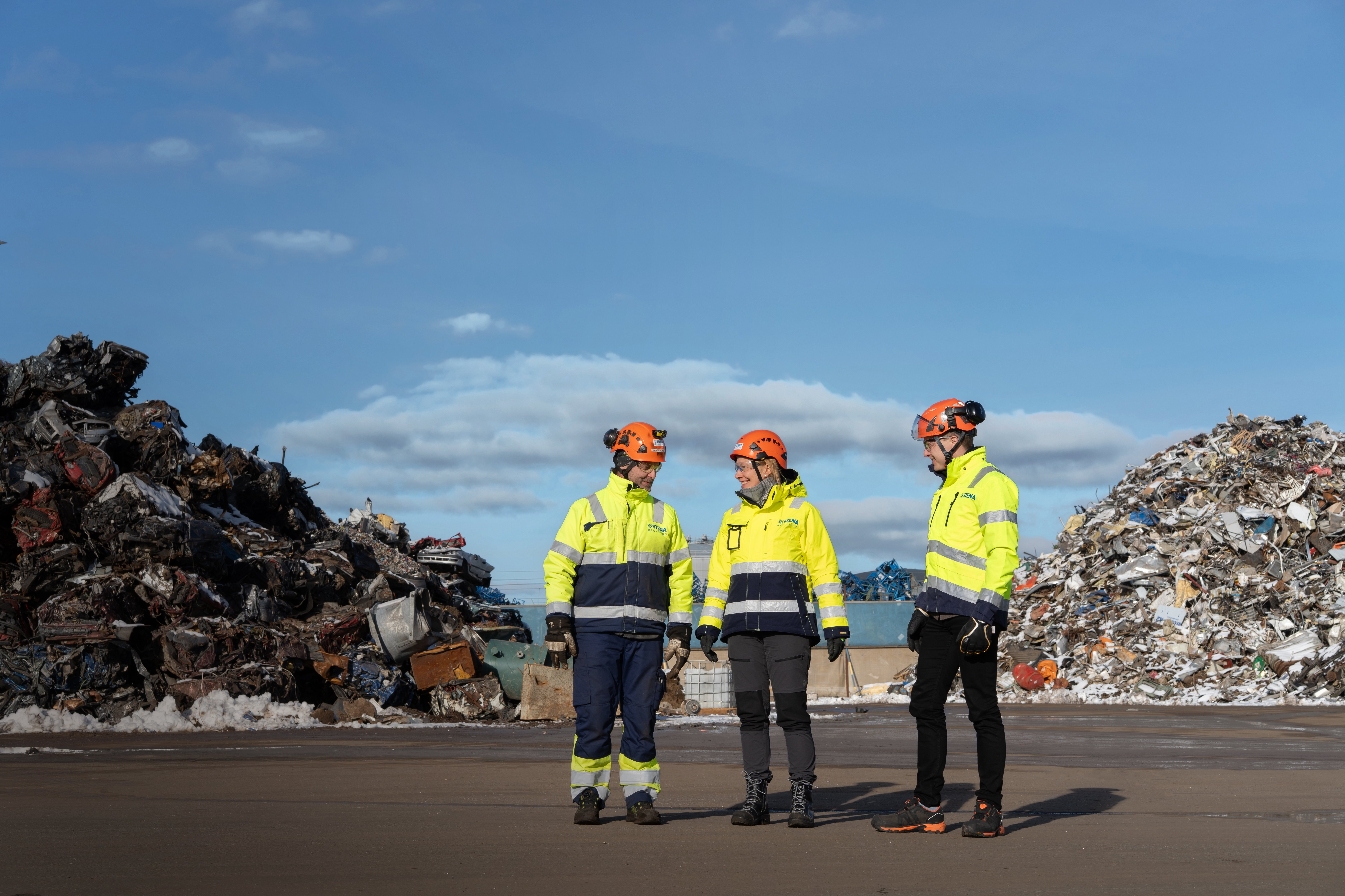 Three Stena Metall employees in hi-vis working gear are standing outside a recycling facility, where metal scrap is treated and recycled