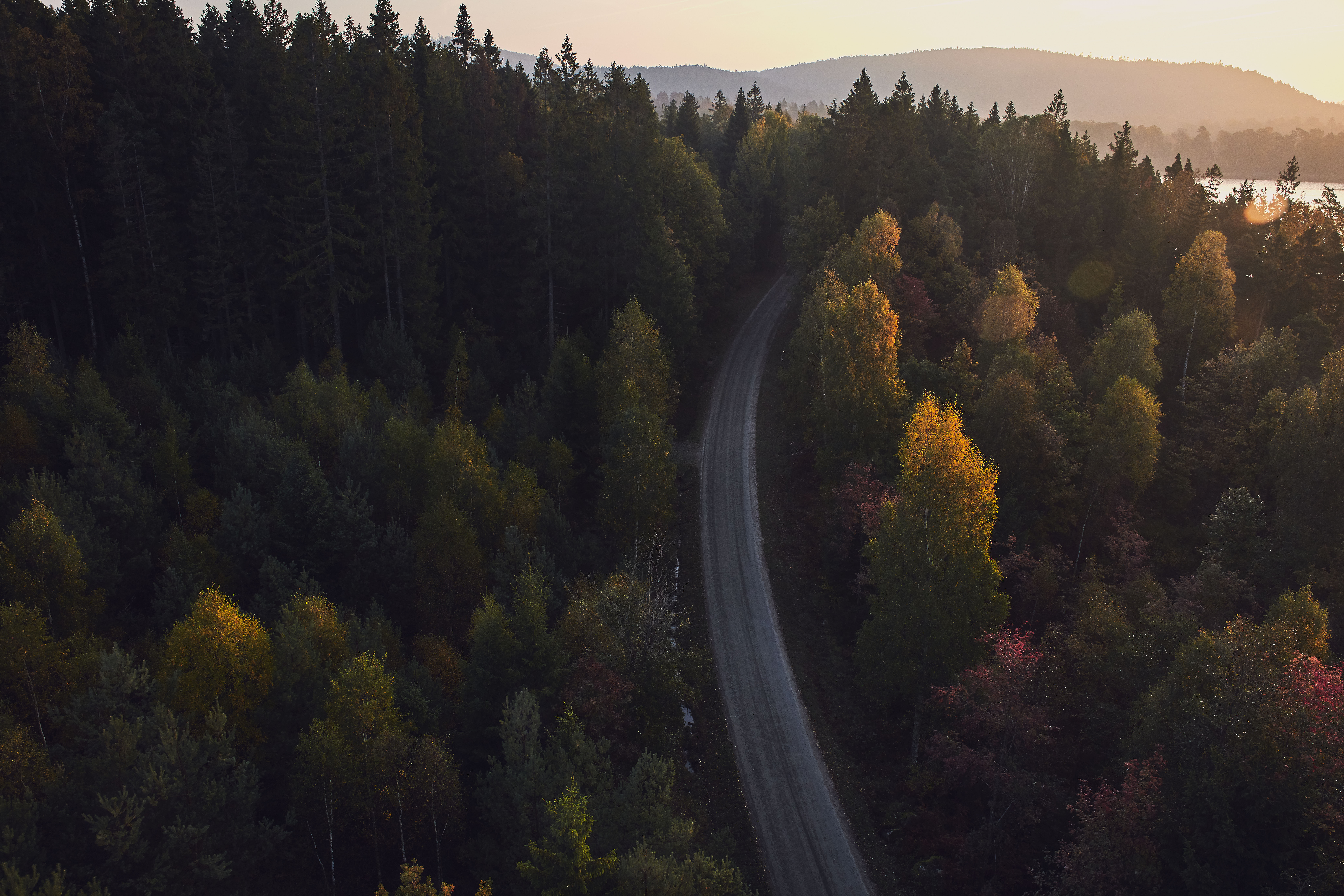 A country-side road seen from above, surrounded by forest. Mountains and water are seen in the background.