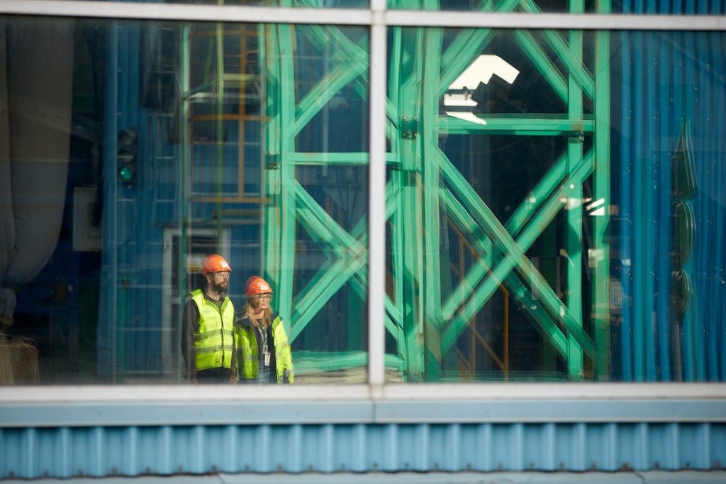 The sight of a male and a female Stena Recycling employee in hi-vis protective gear that stand in a production facility is reflected in a window.