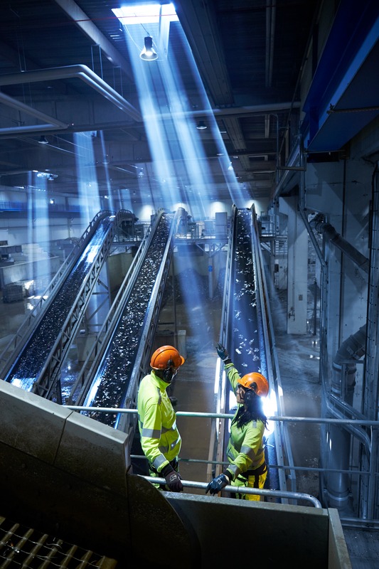 The back of one male and one female Stena Metall employee in hi-vis protective gear and helmet. They stand above and look down on a conveyor belt with scrap metal parts. Sun rays are visible from a window in the ceiling.